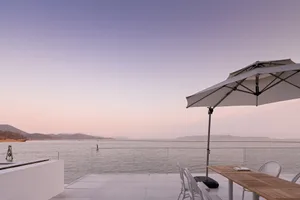 Modern dining area featuring a glass table and chairs, overlooking a serene lake at sunset.