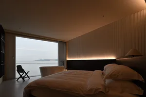 Modern minimalist bedroom with large window overlooking water, featuring textured canvas walls and soft, warm lighting.