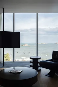 A minimalist residential lounge featuring a large window with a sea view, dark furniture, and round tables.