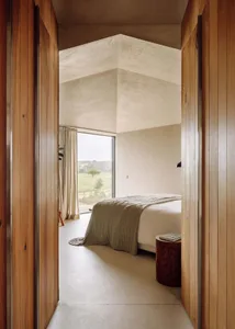 A minimalist bedroom featuring a bed with a wooden frame and a concrete floor, with a view of a natural landscape through a l