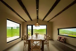 Minimalist dining room featuring a wooden table and chairs, large windows overlooking green fields, and rustic wooden beams o