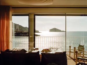 An elegant living room features glass walls overlooking the ocean, with concrete flooring and wooden furniture.