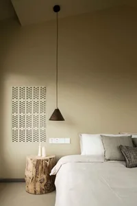 A minimalist bedroom featuring a wooden stump nightstand with a single candle holder and a modern pendant light hanging above