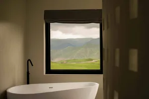 A modern bathroom featuring a white freestanding bathtub with a view of green hills and mountains through a large window with