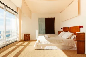 Minimalist bedroom with a bed, wooden headboard, and side tables, featuring a large window with a view of the ocean.