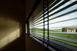 A view of a modern interior room through closed window blinds, showing a peaceful outdoor scene with a wooden structure and g