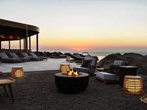 Outdoor lounge with concrete seating, wooden chairs, and wicker stools, overlooking the ocean during sunset.
