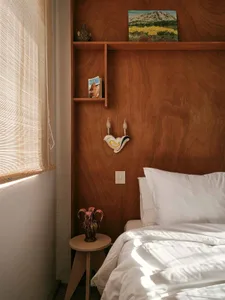 A minimalist bedroom features a bed with white bedding, a wooden headboard, and a wall-mounted sconce. The room is adorned wi