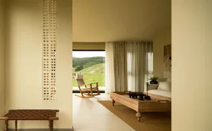 A minimalist living room featuring a wooden sofa and a view of green hills outside.