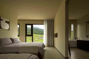 A modern minimalist bedroom featuring neutral-toned decor, a cozy bed, and a floor-to-ceiling window overlooking a green land