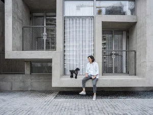 A man sits on a concrete ledge with his black dog in front of a large glass window in a modern apartment building.