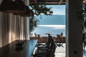 A minimalist patio overlooking the sea, featuring a wooden dining table, metal chairs, and a glass surface reflecting sunligh
