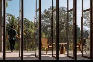 A minimalist residential balcony with wooden furniture and a forest view through large glass doors.