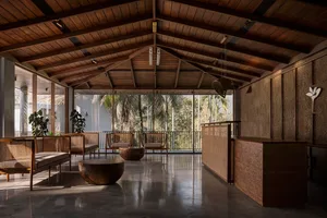 A modern minimalist lobby with wooden furniture, concrete flooring, and a view of palm trees through large windows.