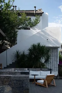 A modern minimalist patio features a white couch, a wooden chair, and tropical plants.