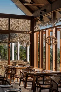A restaurant interior with natural wood furniture and large windows showcasing a tropical outdoor setting.