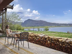 A woman sits at a table near a stacked woodpile by a lake, enjoying the serene outdoor setting.