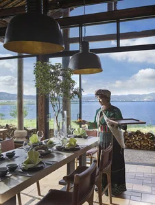 A restaurant interior featuring a wooden dining table, glass windows overlooking a lake, and a person wearing traditional clo