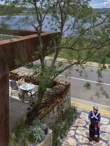 A cozy outdoor seating area with a small table and chairs by a wall made of stacked wood logs, near a road and water body, fe