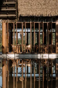 A rustic restaurant with a thatched roof, open folding doors, wooden furniture, and a scenic water reflection.