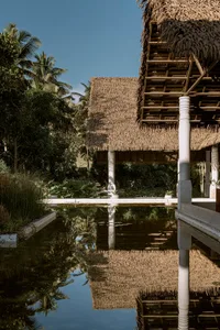 Thatched-roof huts reflected in calm pool water, surrounded by lush tropical foliage and palm trees.