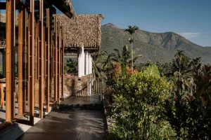 A residential exterior with a thatched roof, wooden columns, and lush greenery against a mountainous backdrop.