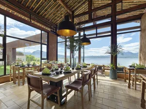A tropical restaurant interior with a wooden floor, large windows overlooking the water, and stylish decor featuring greenery