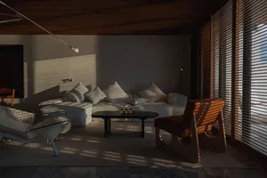 A minimalist living room with a white couch, wooden chairs, and a black table, featuring window blinds casting shadows on the
