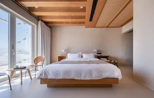 A minimalist bedroom featuring a wooden platform bed with white linens, white bedside lamps, and a coffee table with a drink