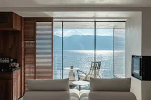 Contemporary living room overlooking the sea with wooden shutters, glass walls, and concrete furniture.