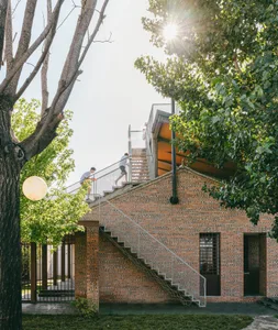 A modern brick house with sunlight shining through trees and a metal staircase leading up to the second floor.