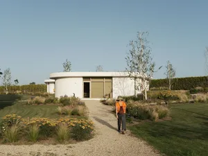 A person wearing an orange shirt and black pants walks towards a circular modern home with concrete walls and glass doors.