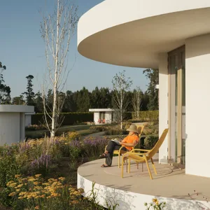 A modern residential exterior with a curved concrete wall and a person sitting on a yellow chair reading a book.