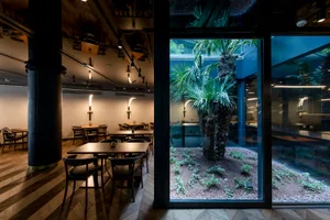 A modern restaurant interior featuring wooden furniture, glass walls, and a central potted plant.