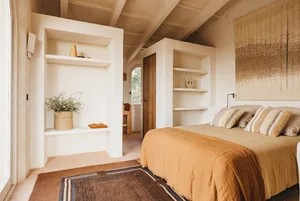 Minimalist bedroom with textured materials, featuring a bed with a yellow blanket, a rug, and built-in shelves.