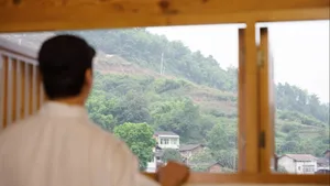 Man standing at window overlooking a mountain landscape with green hills and scattered houses.