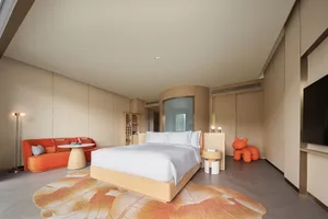 A modern minimalist bedroom featuring a bed with a wooden frame, white bedding, and an orange couch with pillows under a beig