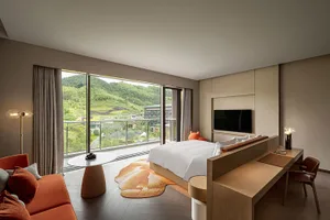 A modern residential bedroom featuring a glass window, a large bed, a wooden desk, and an orange lounge chair.