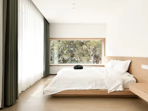 A minimalist bedroom with a bed frame made of light-colored wood and white bedding, framed by large windows with floor-to-cei