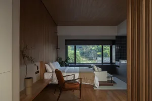 A minimalist bedroom featuring a wood-paneled wall, a bed with white linens, a white armchair, and a view of greenery outside