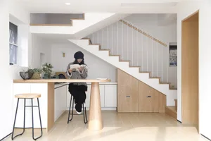 A modern kitchen with white oak wood cabinets, a white countertop, and a white staircase in the background. A woman is sittin