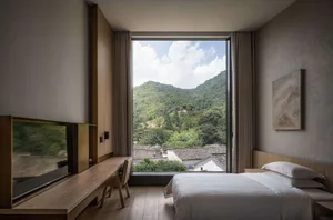 A minimalist bedroom with white furniture and soft textures, featuring a large window overlooking green mountains and rooftop