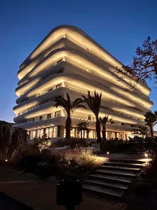 A modern, stacked building illuminated with lights at dusk with palm trees in the foreground.