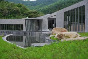 A modern building made of concrete with a pool in the foreground, surrounded by greenery and large rocks.