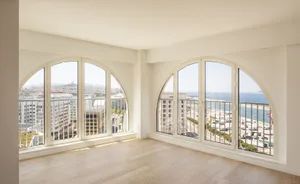 Modern apartment interior with large windows overlooking a cityscape and the ocean, featuring hardwood flooring and large arc