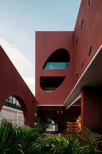 A modern building with a red concrete exterior featuring irregular window cuts and green foliage.