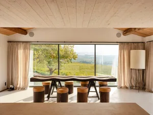 Modern dining room with minimalist design featuring a wooden table and stools, large window overlooking a scenic vineyard.