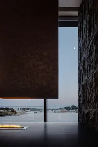 A modern minimalist living room featuring concrete and rock walls, with a minimalist fireplace lit and a view of the outdoor 