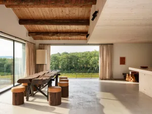 A modern minimalist living room with wooden accents and furniture, featuring large windows overlooking a forested area.
