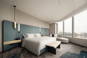 A modern hotel room featuring a bed with a white comforter and a blue headboard, overlooking a cityscape through a large wind